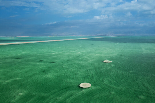 Aerial view of the shimmering turquoise waters of the Dead Sea, revealing intricate salt patterns and circular formations under a dramatic sky, Ein Bokek, Southern District, Israel.