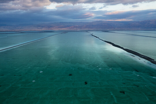 Aerial view of shimmering turquoise salt ponds stretching towards distant rugged mountains under a dramatic cloudy sky, Sdom, Israel.