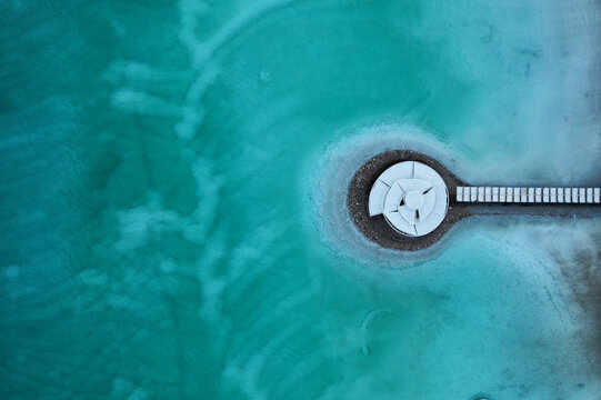 Aerial view of a stark white geometric structure on a circular island, connected by a pier, amidst swirling turquoise and teal water, Ein Bokek, Israel.