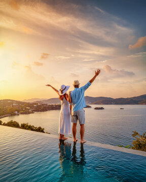 A happy couple on summer vacation standing by the swimming pool enjoys a beautiful sunset behind the sea