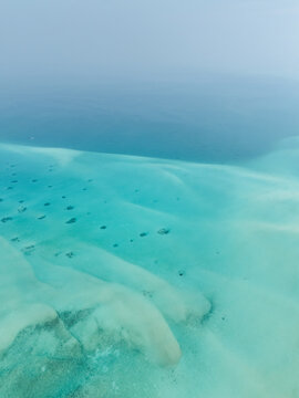 Aerial view of vibrant turquoise shallows revealing sandy patterns and dark coral heads, transitioning into the deep, hazy blues of the ocean horizon, Thuraakunu, Thiladhunmathee Uthuruburi, Maldives.