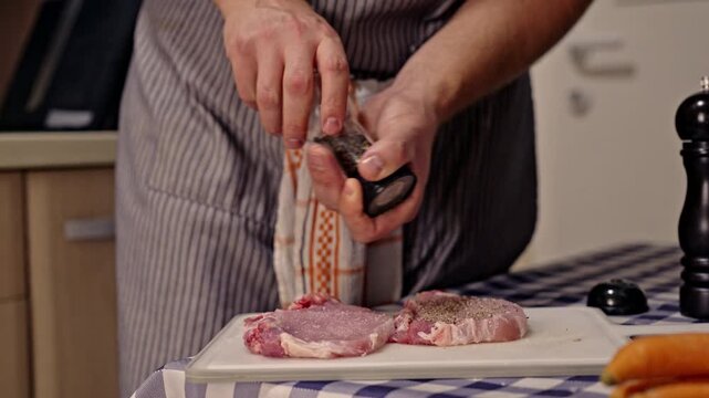 A person in a striped apron grinds fresh black pepper onto two raw meat cuts on a white cutting board. This culinary preparation highlights the essential steps of seasoning before cooking a meal.