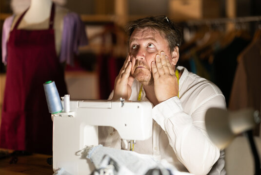 Overworked middle-aged male tailor sitting on table next to sewing machine