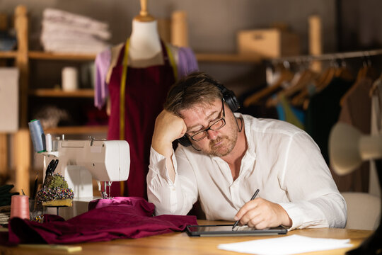 Middle-aged man wearing earphones drawing on tablet in sewing workshop at night