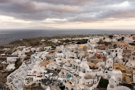 Aerial view of white buildings cascading down the cliffs towards the sea under a brooding sky, Oia, Thira, Greece.
