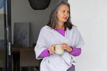 Naklejka premium Senior woman standing near sliding glass door holding ceramic mug, wearing magenta top and wrap