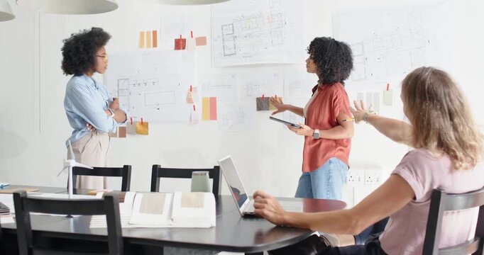 Diverse female team presenting with tablet samples pointing at wall plans deciding colors at studio