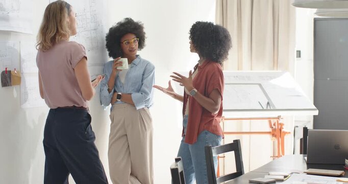 Diverse female team reviewing pinned sketches at studio with tablet, right woman leading discussion