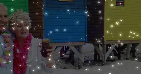 Fototapeta premium Standing couple wearing pink, rainbow leis holding tiki glass by colorful huts on sand, copy space