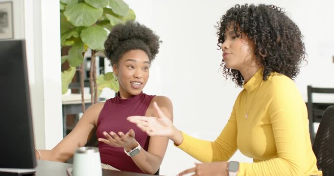 Diverse female coworkers left woman gesturing at monitor, right woman pointing and guiding at desk