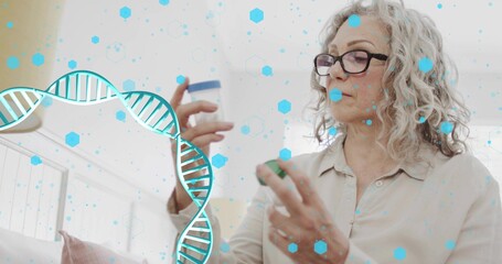 Reading senior woman in beige blouse, black glasses holding pill bottles on sofa with DNA graphics © vectorfusionart