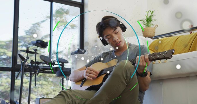 Strumming teen Korean playing guitar in living room by windows, with drum kit headphones overlays