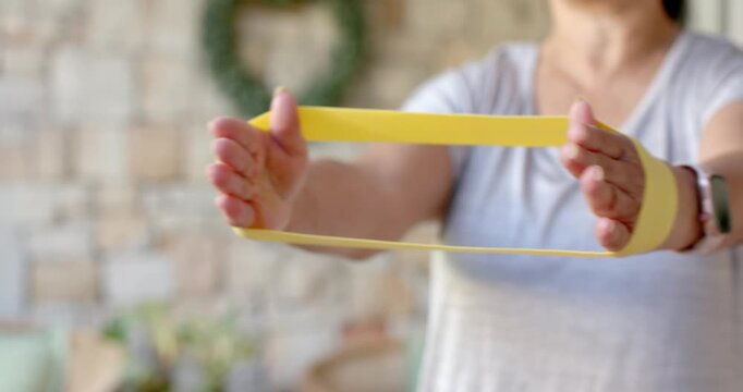 Senior Asian female forming loop with yellow band on porch wearing smartwatch, pulling for rehab