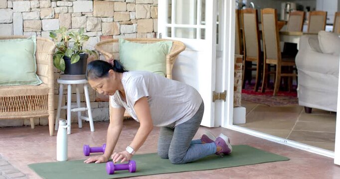 Senior woman kneeling on green patio mat, pulling purple dumbbell up in leggings for fitness