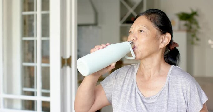 Senior Asian woman in gray T-shirt picking up mint bottle by glass door, drinking for hydration