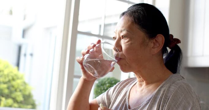 Senior Asian woman at kitchen window pausing to take pill, lifting glass, sipping, swallowing water