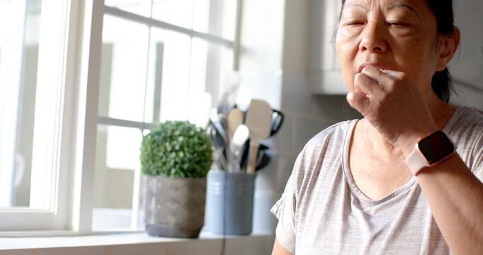 Senior Asian woman in kitchen prompted by bottles placing pill in mouth, lifting glass, swallowing