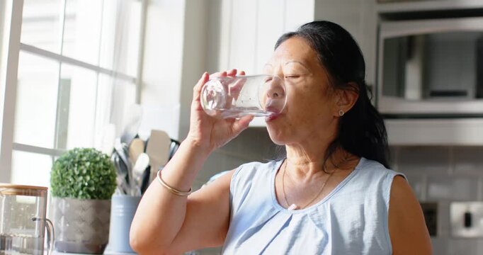 Senior Asian woman lifting glass, sipping water, inspecting remaining liquid by kitchen window