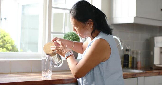 Senior Asian woman in kitchen lifting pitcher removing wooden lid filling empty glass