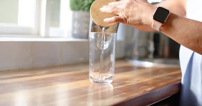 Senior Asian woman placing glass at wood counter, tilting pitcher pouring to fill glass, copy space