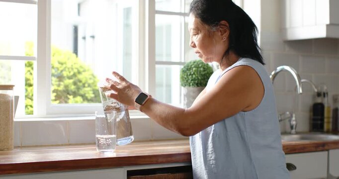 Senior Asian woman thirsty, lifting lid, pouring water from pitcher into glass to drink at counter