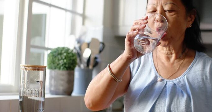 Reaching for glass at home counter, Senior Asian woman drinking, gazing out window, copy space