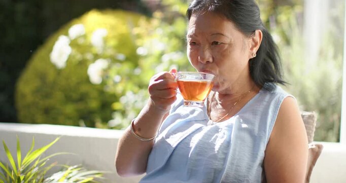 Senior Asian woman sitting on patio chair camera zooming lifting teacup inhaling and sipping tea
