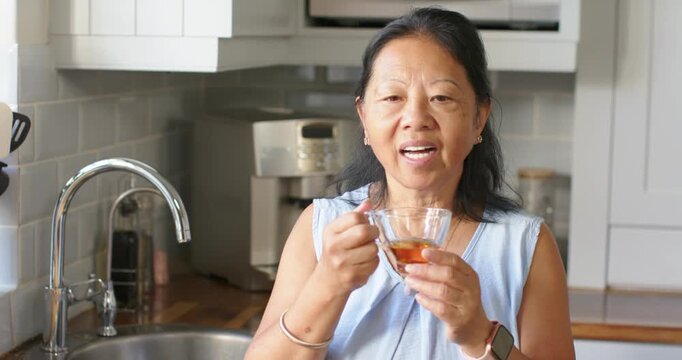 Senior Chinese woman smelling tea aroma, closing eyes, sipping to taste from glass cup at sink