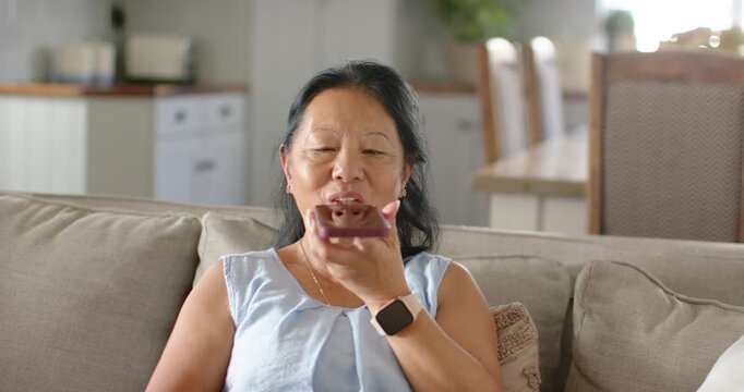 Asian senior woman raising mauve phone recording voice note on sofa at home wearing rose-gold watch