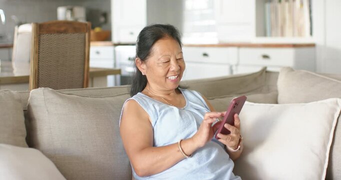 Senior Asian woman starting smiling, tapping and scrolling pink phone on sofa in light blue top