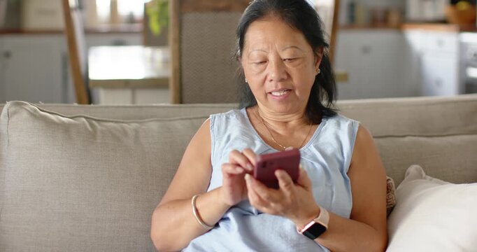 Senior Asian woman holding and swiping pink phone while reading texts on sofa wearing blue top
