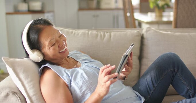 Asian Senior woman reclining on sofa wearing headphones, hearing audio, reacting by tapping tablet