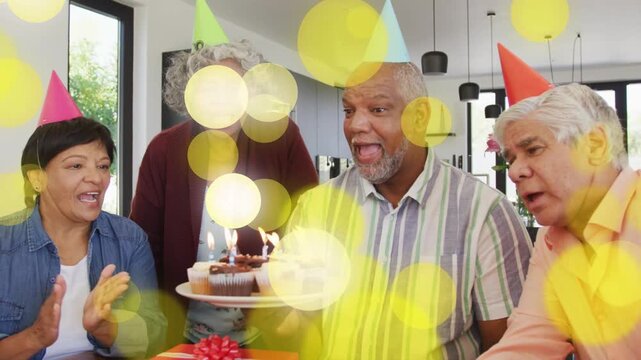 Man holding cupcakes blowing out candles after group singing for birthday, yellow bokeh crossing