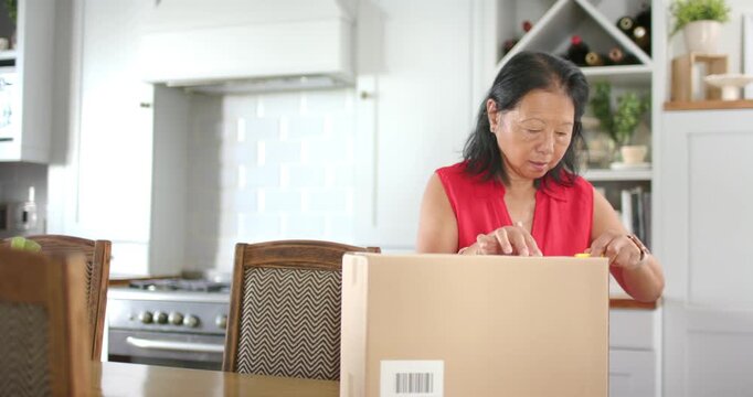 Senior Asian woman inspecting box at kitchen table using yellow cutter opening box viewing contents