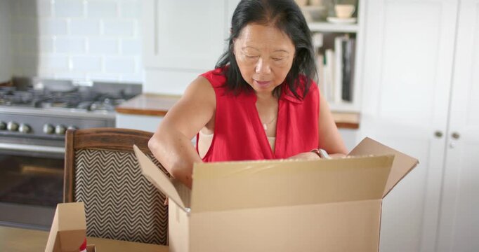 Senior Chinese female opening large cardboard box at kitchen table reaching and inspecting contents
