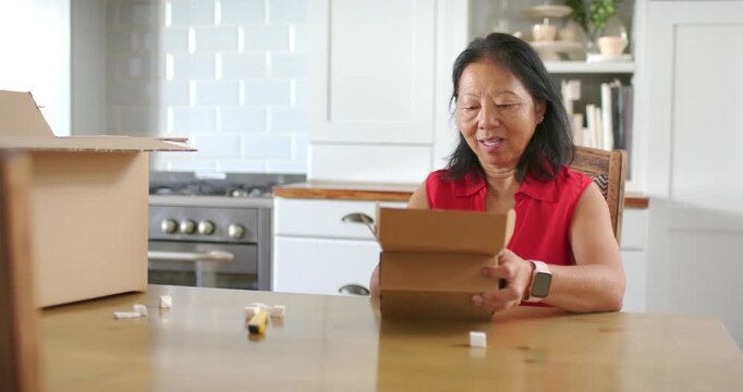 Senior Asian woman in red blouse opening delivery box at kitchen table inspecting white bottle