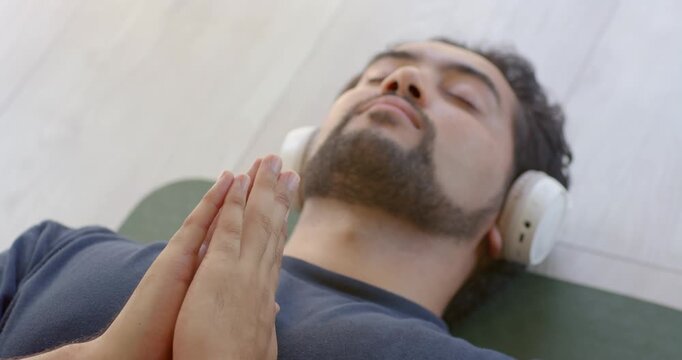 African man lying on green mat wearing white headphones praying hands breathing causing faint rise