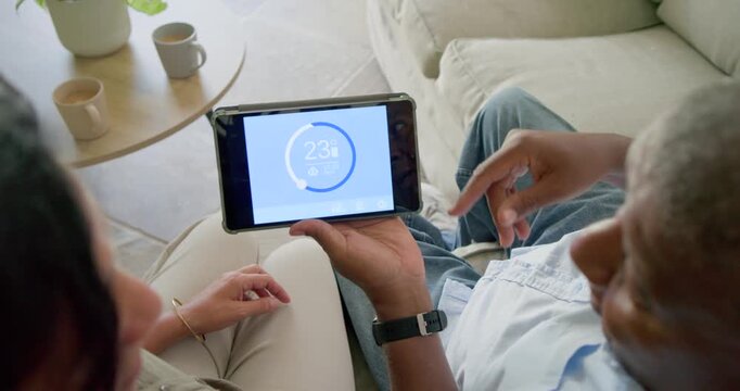 Man is pointing at tablet, woman is leaning and adjusting thermostat on sofa by coffee table