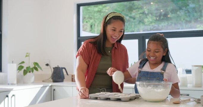 Asian mom holding tin, guiding African-American schoolgirl ladling batter in kitchen making muffins