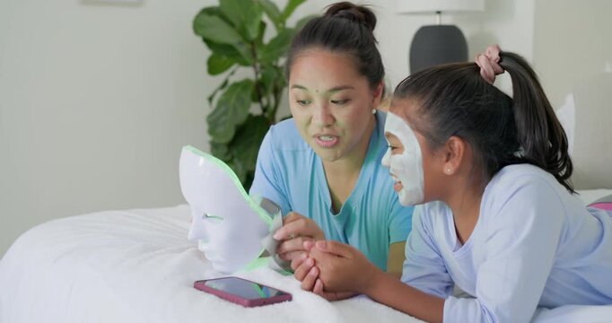 Asian mother demonstrating LED mask on bed with African American teen daughter wearing sheet mask