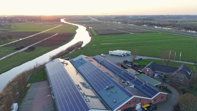 Sunset Aerial Showing Solar Panels On Barn Roofs, Parked Service Vehicles, Winding River And Surrounding Fields Modern Renewable Energy Installation Framed By Warm Light And Rural Context.