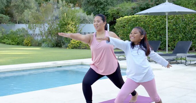 Asian mother stepping into lunge, African American girl mirroring, doing yoga on purple mat by pool