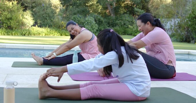 Diverse female family stretching on pool mats, laughing making teen look at water bottle for warmup
