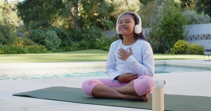 African American teen in leggings sitting on mat by pool doing breathwork via headphones for calm