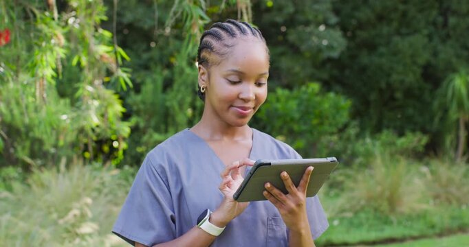 African woman in gray scrubs standing in grassy park, tapping tablet, smiling, checking smartwatch