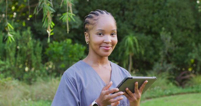 Female clinician in scrubs in garden, tapping tablet after alert, checking notes, smiling