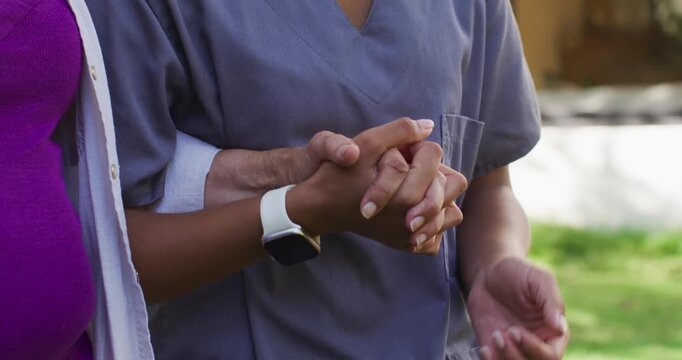 African American senior woman clasping hands around adult woman in scrub on lawn, comforting, watch