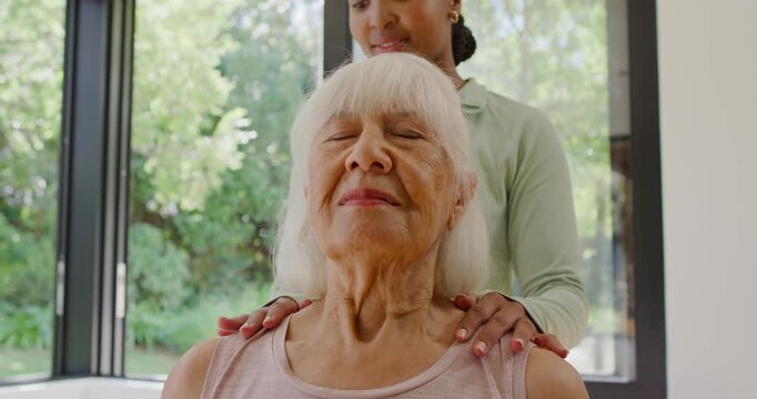 Female aide placing hands on senior in pink top, massaging shoulders for calm at window