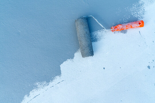 Paint roller with orange handle applying blue paint on a textured white concrete surface. Renovation, home improvement, and construction concept showing professional wall painting progress.