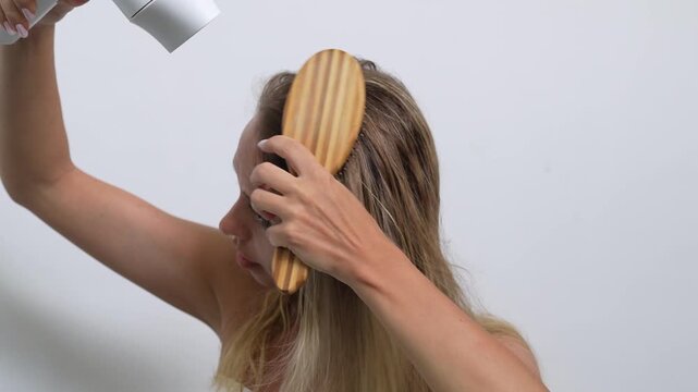 Young woman blow drying and brushing her hair at home. Daily haircare routine. Styling, grooming and beauty care concept with natural blonde hair